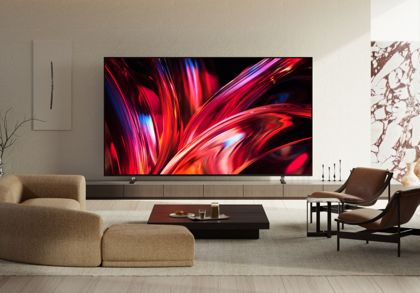Wider view of a modern living room with a wall-mounted TV displaying films like GOAT and Anaconda. A slim soundbar sits below the screen, with a beige sofa, coffee table, and chairs completing the setup.