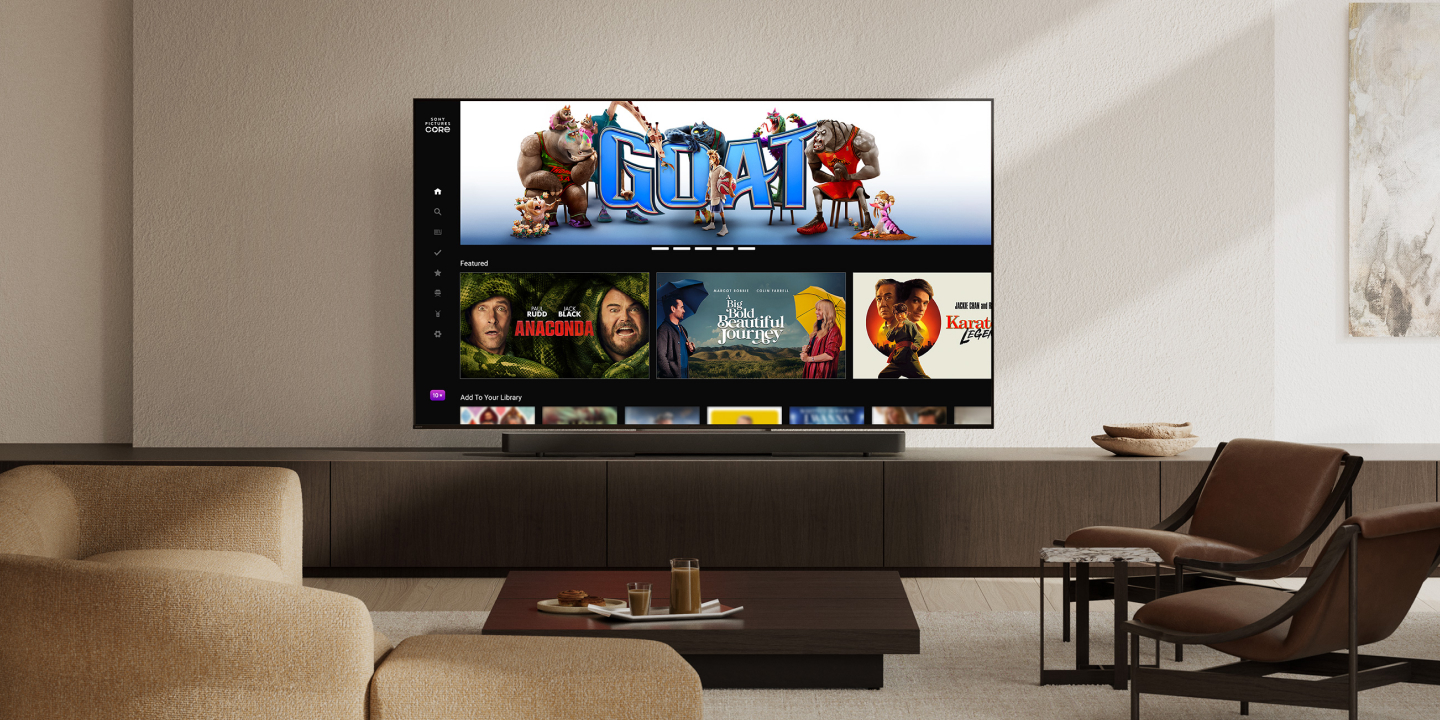 Wider view of a modern living room with a wall-mounted TV displaying films like GOAT and Anaconda. A slim soundbar sits below the screen, with a beige sofa, coffee table, and chairs completing the setup.