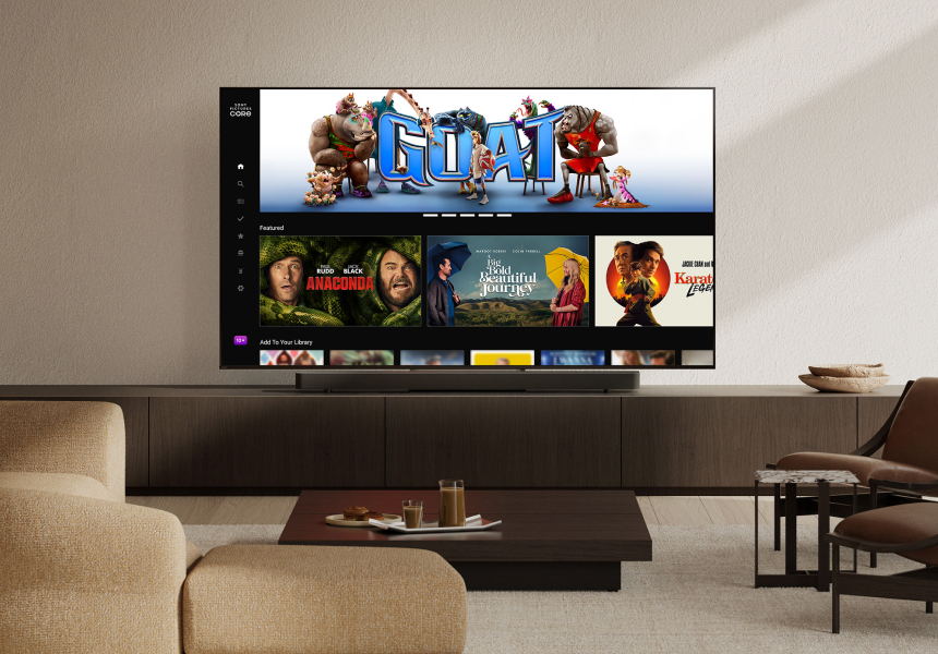 Wider view of a modern living room with a wall-mounted TV displaying films like GOAT and Anaconda. A slim soundbar sits below the screen, with a beige sofa, coffee table, and chairs completing the setup.