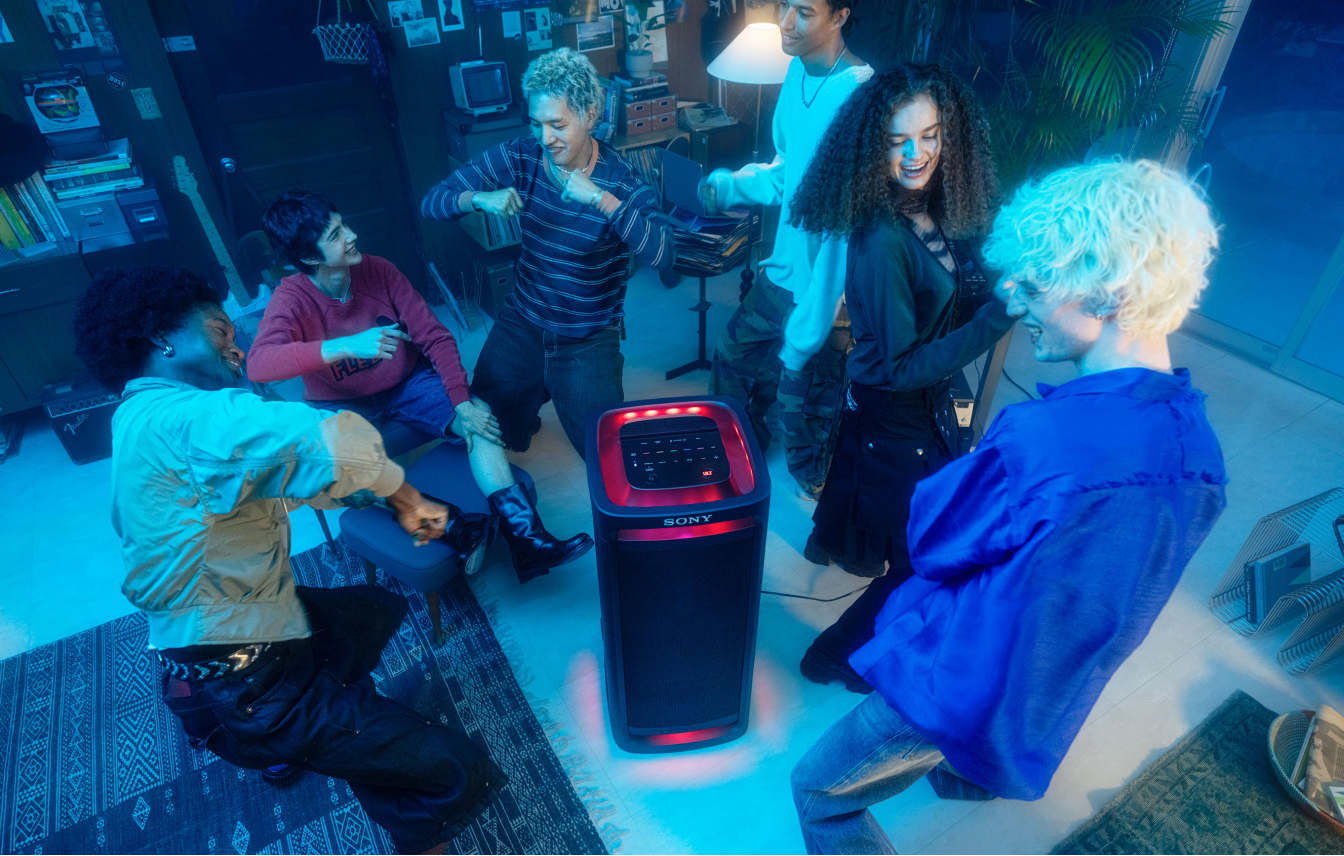 A group of people dancing in a darkened room around the TOWER 9AC speaker with its party lights on